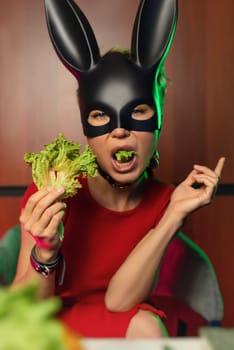 A beautiful girl in a bdsm rabbit mask and a bright red dress eats lettuce leaves promoting a healthy lifestyle and vegetarianism