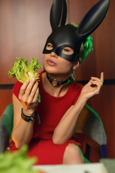 A beautiful girl in a bdsm rabbit mask and a bright red dress eats lettuce leaves promoting a healthy lifestyle and vegetarianism