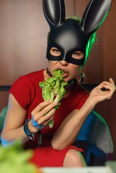 A beautiful girl in a bdsm rabbit mask and a bright red dress eats lettuce leaves promoting a healthy lifestyle and vegetarianism