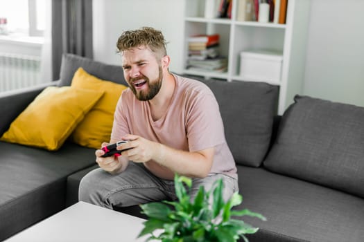 Young man spending time at home, sitting on a couch in apartment and playing arcade car video games on console. Male using controller to play street racing drift simulator