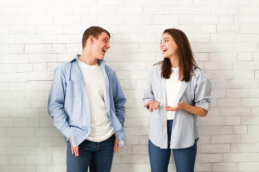 A guy and a girl are standing together in front of a brick wall, looking towards the camera with neutral expressions. Both are dressed casually.