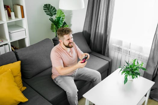 Young man spending time at home, sitting on a couch in apartment and playing arcade car video games on console. Male using controller to play street racing drift simulator