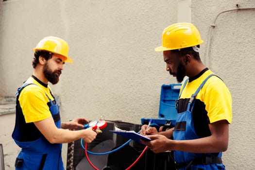 Trained engineer using set of manometers to read pressure of liquids and gases in condenser unit while expert repairman writes air conditioner faulty components report list on clipboard