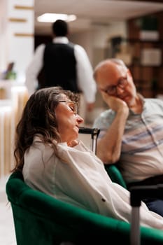 Detailed shot of retired senior woman and man, each sleeping on cozy couch while waiting to register for hotel room reservation. Photo focus on elderly female tourist resting in exclusive lounge area.