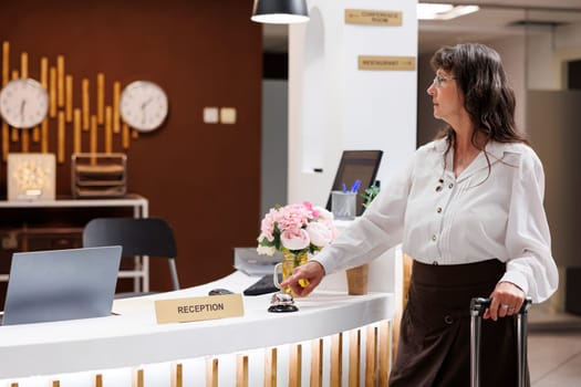Elderly retired woman approaches the front desk and rings the hotel silver service bell to ask for help. Senior female visitor with suitcase awaits help from resort staff at registration counter.