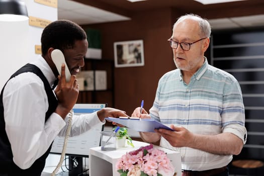 African American concierge answers the telephone, caucasian retired old man signs reservation forms at hotel front desk. While receptionist is on phone call, elderly customer fills out check-in forms.