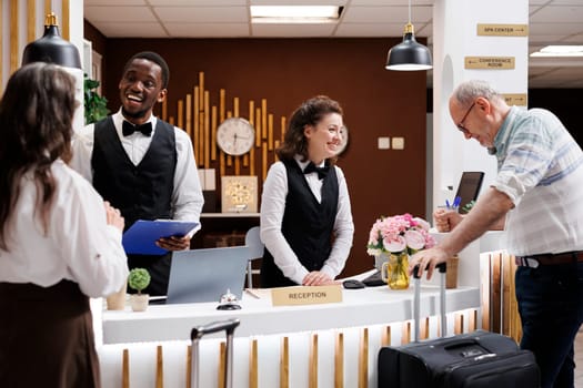 Retired senior couple arrive at hotel, assisted by smiling multiethnic staff at front desk. Check-in, paperwork, luggage. Luxury hotel lobby bustling with tourists. Excited for vacation.