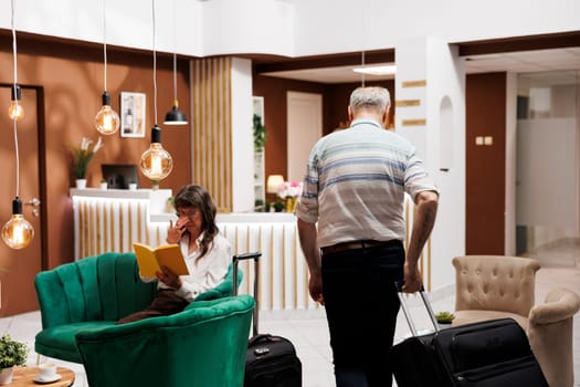 Arriving in contemporary hotel lobby is an elderly caucasian man who goes to front desk to check in. Senior guy tourist carrying luggage as he approaches registration desk to make hotel reservations.