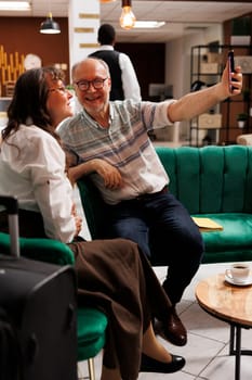 Elderly retired couple using smartphone to take photos in foyer of elegant hotel. Senior old husband and wife photographing enjoyable holiday at luxury resort while keeping their baggage close by.