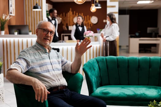 Portrait shot of old man seated on cozy sofa in exclusive hotel reception, waving at camera. Comfortable lobby offers relaxing lounge area for elderly male traveler waiting for check-in procedure.