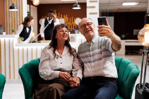 Retired senior couple videocalling family with smartphone from hotel reception. Smiling elderly male and female tourists enjoying vacation in lobby. Professional receptionists working in background.