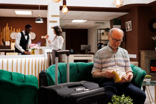 Speaking to young concierge at main desk of luxury resort is caucasian female traveller. While old woman completes booking process at counter an elderly man reading a book relaxes in hotel lobby.
