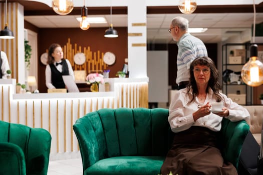 Elderly caucasian woman seated on sofa with cup of coffee at luxurious lounge area for check-in. Senior man arrives in hotel lobby while old female tourist waits on couch for booking procedure.