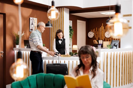 Receptionist helps senior man traveller check in when he arrives at hotel front desk. While elderly woman unwinds in elegant lounge area female concierge assisting male tourists with accommodation.