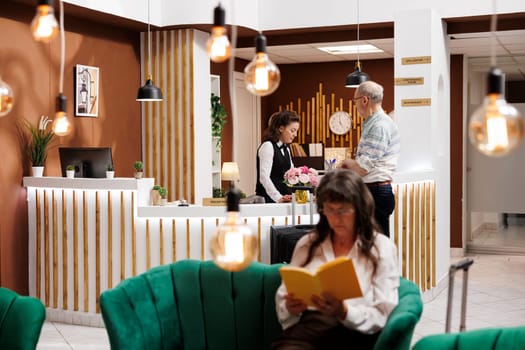 Elderly male guest arrives at front desk of hotel lobby, greeted by friendly female receptionist. Senior woman in cozy lounge area sitting on comfy sofa and reading a book while waiting for check-in.