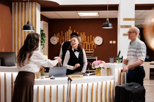 In hotel reception, elderly male and female travelers at registration counter awaiting reservation procedure. Retired senior woman giving personal documents to concierge for check-in.