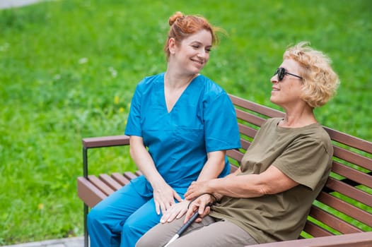 A nurse and an elderly blind woman are sitting on a bench in the park