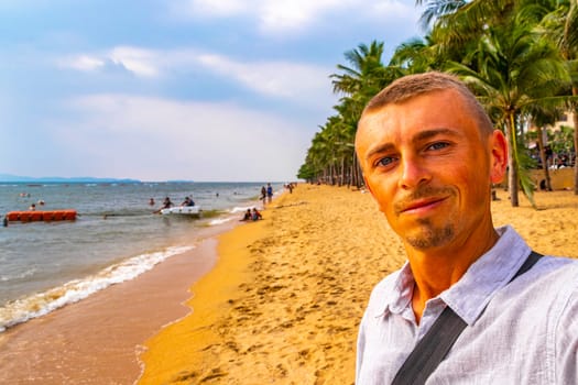 Tourist man takes selfie photo at tropical beach waves water sand people boat palm trees and skyscrapers in Jomtien Beach Pattaya Bang Lamung Chon Buri Thailand in Southeastasia Asia.