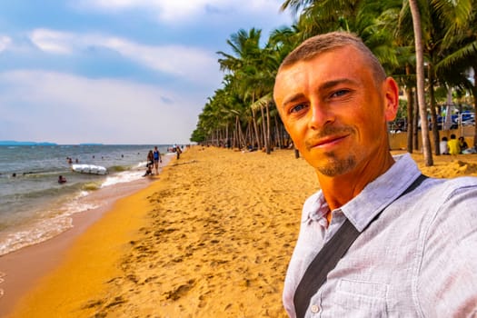 Tourist man takes selfie photo at tropical beach waves water sand people boat palm trees and skyscrapers in Jomtien Beach Pattaya Bang Lamung Chon Buri Thailand in Southeastasia Asia.