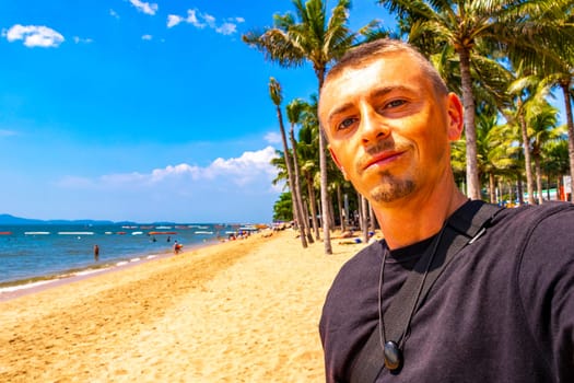 Tourist man takes selfie photo at tropical beach waves water sand people boat palm trees and skyscrapers in Jomtien Beach Pattaya Bang Lamung Chon Buri Thailand in Southeastasia Asia.