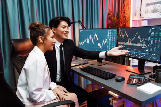 Asian businessman showing dynamic stock exchange currency rate statistic online to share with coworker, explaining to compare with high or low on computer screen at night neon modern office. Infobahn.
