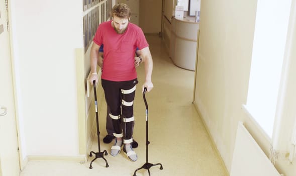 Young disable man in orthosis walking with support of two walking cane in the rehabilitation clinic. Doctor helping him.