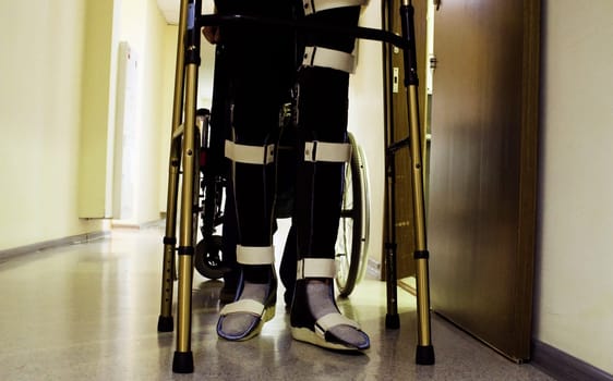 Legs of young disabled man in orthosis walking holding a walking frame through the corridor of the rehabilitation clinic.