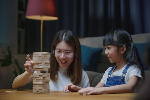 Asian young mother playing game in wood block with her little daughter in home living room at night time, Smiling woman help teach child play build constructor of wooden blocks, education