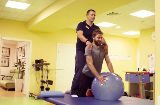 Young disabled man doing exercises at the rehabilitation center with a ball. Doctor physiotherapist helping him. Healthy gymnastics. Active people.