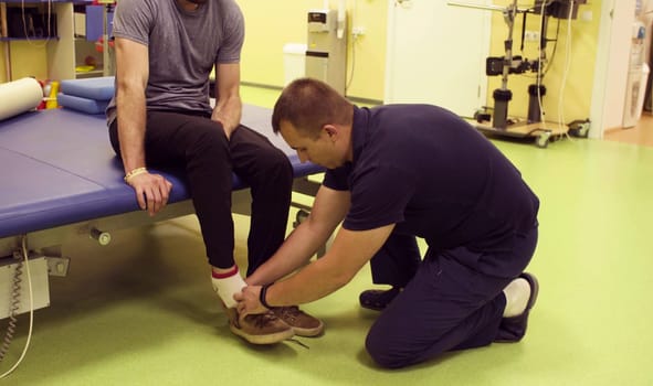 Doctor physiotherapist in the rehabilitation center puts on shoes for the disabled man