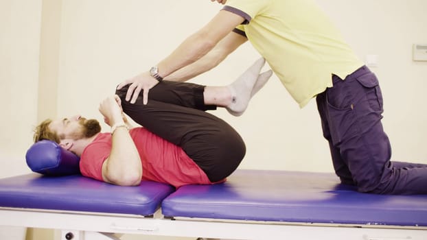 Doctor physiotherapist helping young disabled man, doing leg exercises at the rehabilitation center. Health reductive gymnastics.