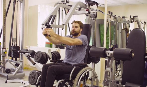 Young disabled man in wheelchair doing strength exercises for hands in the rehabilitation clinic. Doctor physiotherapist helping him