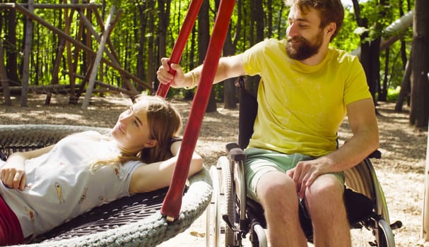 Happy young disable man in a wheelchair in the park with his wife. Woman is lying on a swing and a man is pushing a swing