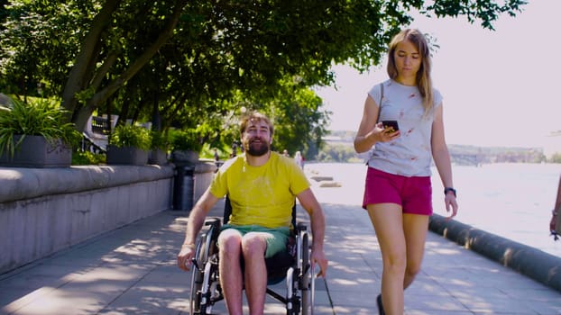 Happy young disable man in a wheelchair in the park with his wife. They are walking along the embankment near the river