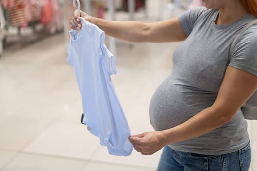 Caucasian pregnant woman chooses baby clothes in a store. Faceless expectant mother in the 3rd trimester