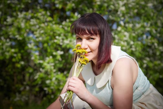 Portrait of beautiful young woman near apple trees and yellow dandelion flower in park on sunny day. Springtime