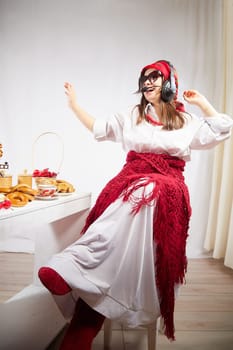 Girl in red scarf and large headphones with microphone having fun at table with delicious food for Orthodox holiday Maslenitsa or Easter. Funny Woman operator, Freelancer, blogger singing and dancing