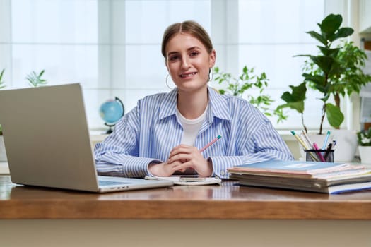 Portrait of high school, college student smiling young female sitting at desk with laptop computer looking at camera. Education, training, e-learning, 16,17,18 year old youth