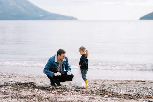 Dad and little girl collecting trash in a bag on the beach by the sea. High quality photo