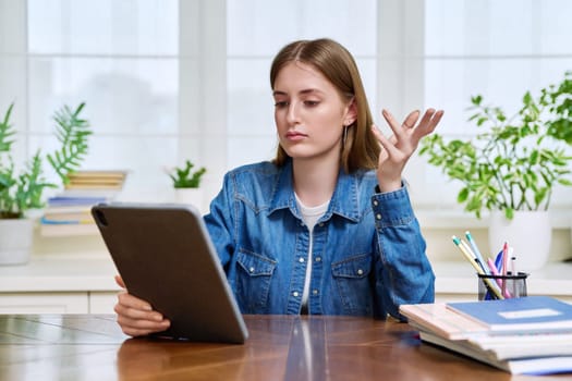 Young teenage female student sitting at home looking talking in web camera of digital tablet. Girl teenager 16,17,18 years old studying remotely, video conference call, e-learning technology education