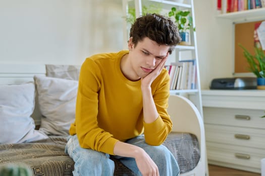 Sad upset tired young man sitting on couch at home, touching his head with hands. Health problems, headaches, troubles, difficulties in study, family relationships, mental health, stress, depression