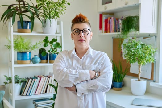 Portrait of smiling confident middle-aged woman in glasses with red haircut, female with crossed arms looking at camera in home interior. Mature people, lifestyle, health, life concept