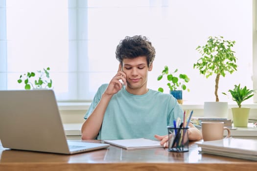 Young guy student talking on the phone, sitting at his desk at home.