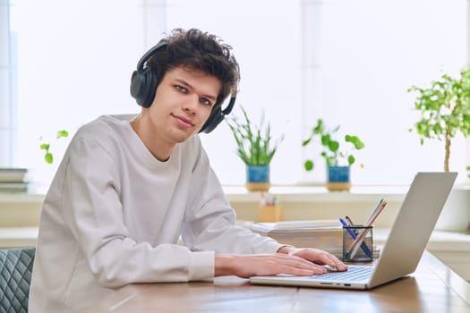 Young college student guy sitting at desk with laptop computer looking at camera. Handsome curly young male wearing headphones in home interior. Education, lifestyle, youth 19,20 years old