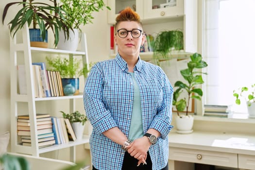 Portrait of smiling middle-aged woman in glasses with red haircut looking at camera in home interior. Mature people, lifestyle, health, life concept