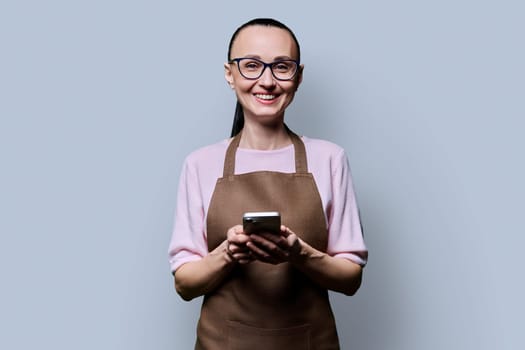 Portrait of 30s woman in apron with smartphone looking at camera on grey background. Smiling female using mobile phone texting receiving sending order. Technologies applications service small business