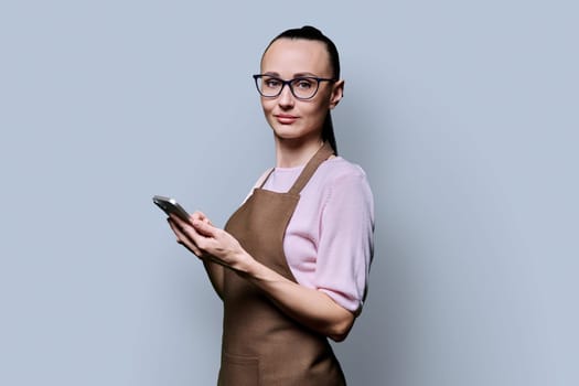 Portrait of 30s woman in apron with smartphone looking at camera on grey background. Serious female using mobile phone texting receiving sending order. Technologies applications service small business