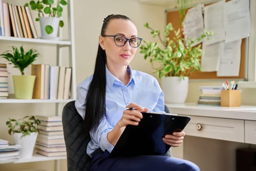 Portrait of smiling female psychotherapist with clipboard at workplace in office. Professional mental therapist counselor psychologist social worker looking at camera. Health care services, treatment