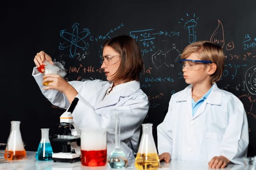 Skilled teacher and student in laboratory wear lab coat stand and experiment about science of chemistry in STEM class. Teacher pour red liquid down to yellow beaker. Table put many flask. Erudition.
