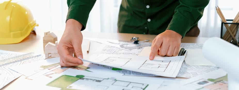 A portrait of professional male engineer shows mistake point in blueprint on messy table with architectural plan and yellow helmet placed at modern office. Closeup. Focus on hand. Delineation.